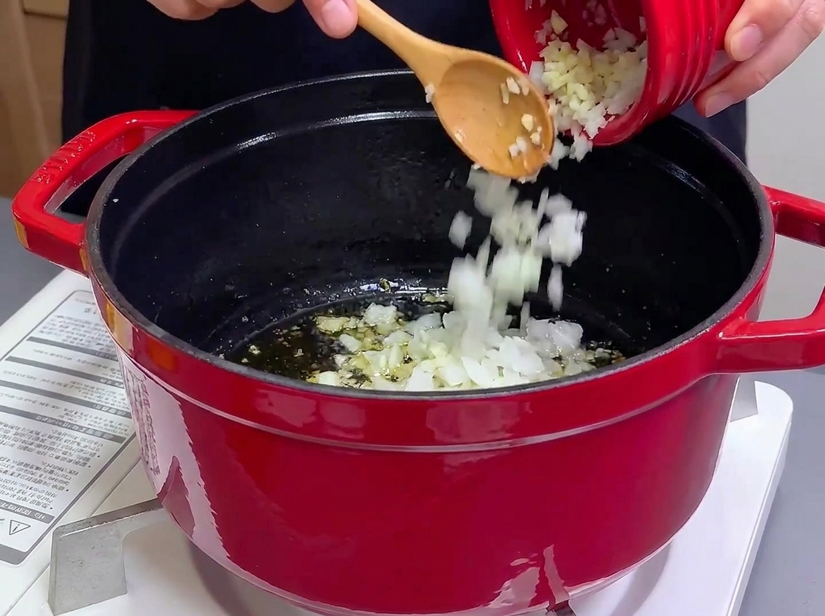 Gehackte Zwiebeln und Knoblauch werden aus einer kleinen roten Schüssel in einen Gusseisentopf mit heißem Öl gegossen.