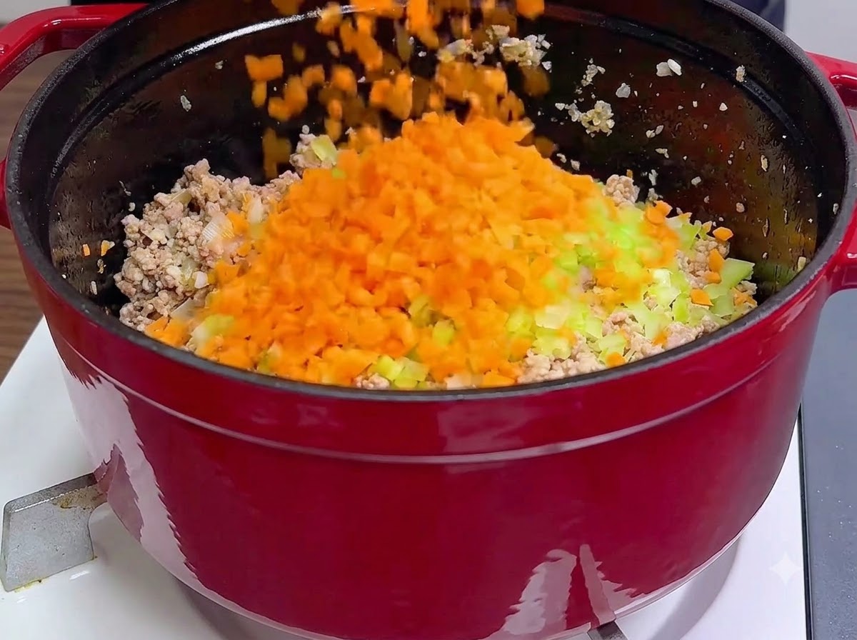 Frisch gewürfelte orangefarbene Karotten und grüner Sellerie werden in einem roten Topf direkt über das gekochte Hackfleisch gegossen.