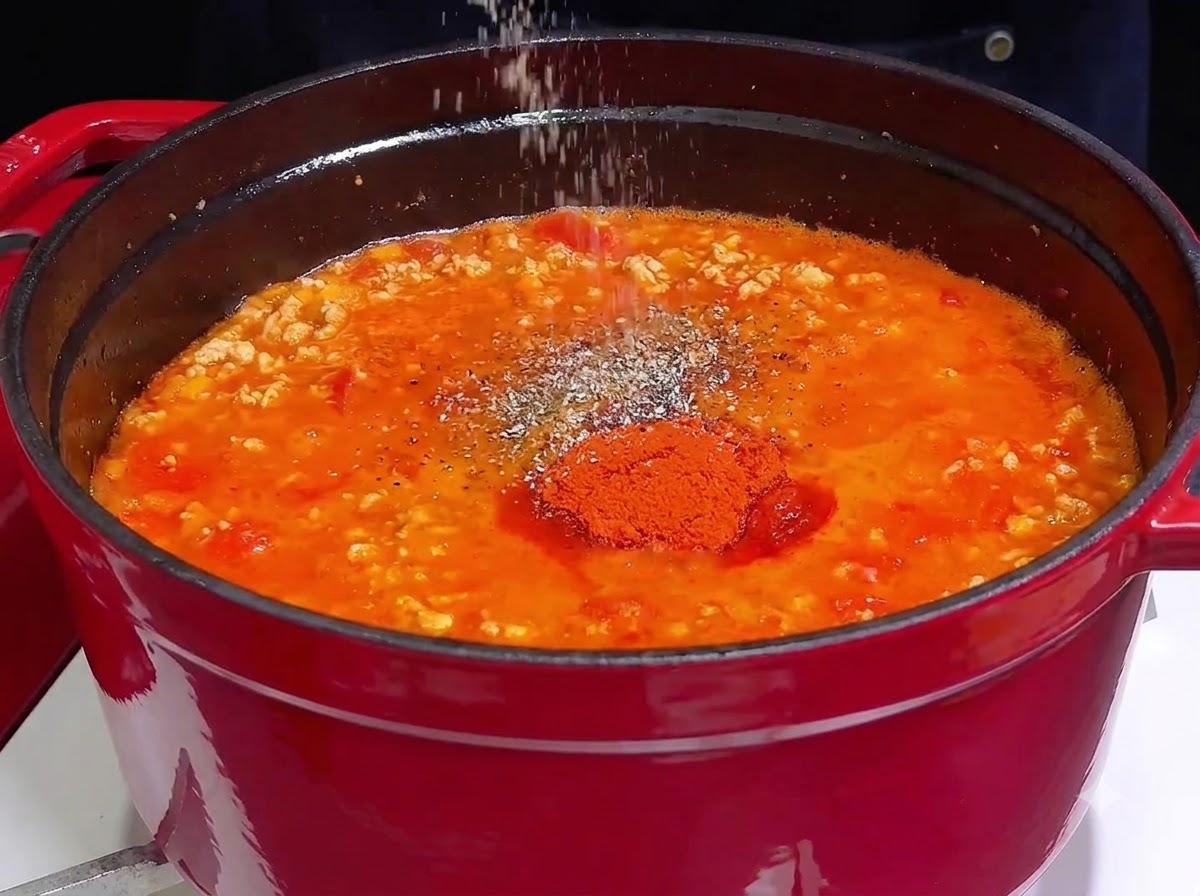 Eine Hand streut Salz und Gewürze über eine leuchtend rote, köchelnde Fleischsauce in einem Gusseisentopf.