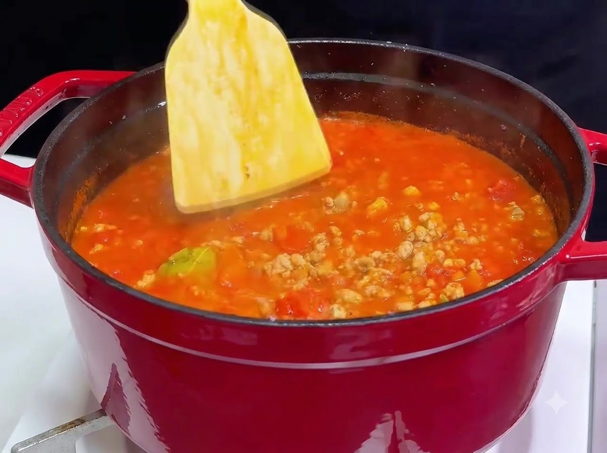 Ein Holzspatel rührt Rotwein in einen roten Gusseisentopf, der mit blubbernder Tomaten-Fleisch-Sauce gefüllt ist.