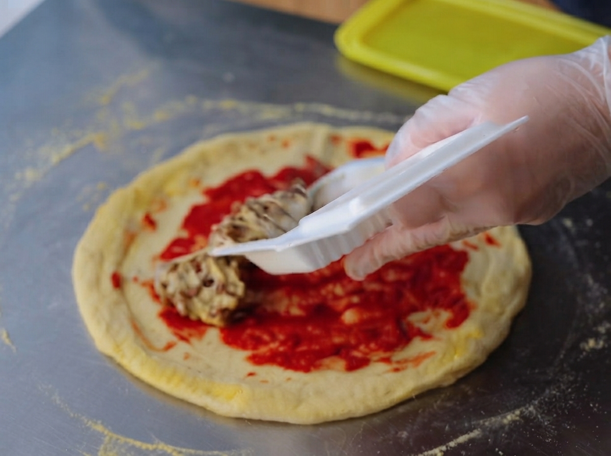 Eine handgeschützte Hand verteilt eine klebrige, fadenartige Natto-Mischung gleichmäßig über der hellroten Tomatenmarkschicht auf einem rohen Pizzateig.