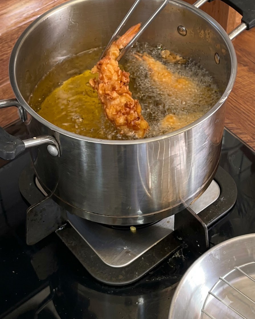 Metallzangen halten eine goldbraune, knusprig frittierte Garnele über einem heißen Öltopf.
