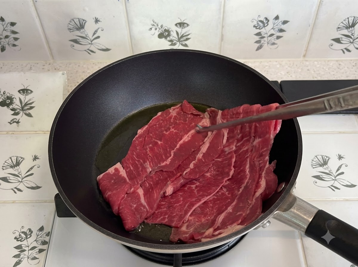 Dünn geschnittenes fettes Rindfleisch wird in einer dunklen Pfanne mit einer Küchenzange angebraten.