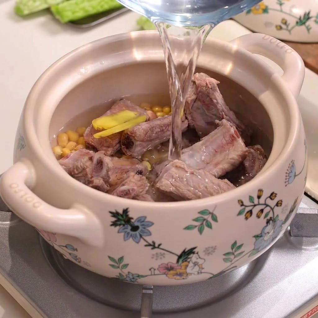 Wasser wird aus einem Krug in einen verzierten Keramiktopf gegossen, der mit Schweinerippchen und Sojabohnen gefüllt ist.