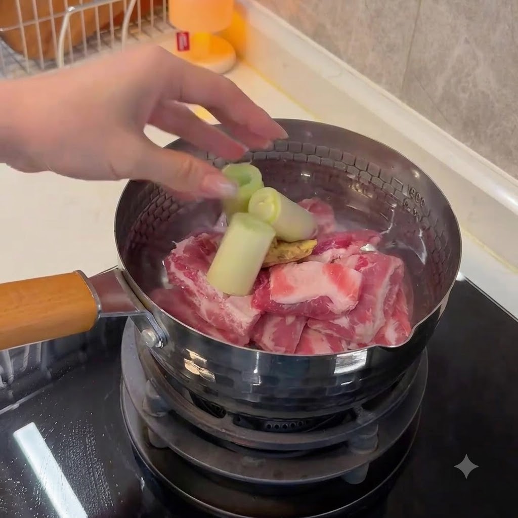 Eine Hand wirft große Stücke Frühlingszwiebel und geschnittenen Ingwer in einen Topf mit Schweinerippchen und Wasser.