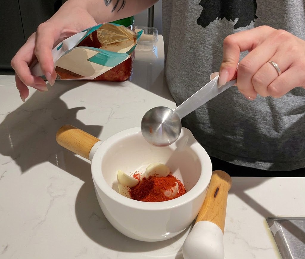 Rotes Chilipulver wird in einen weißen Mörser gefüllt, der Knoblauchzehen enthält.