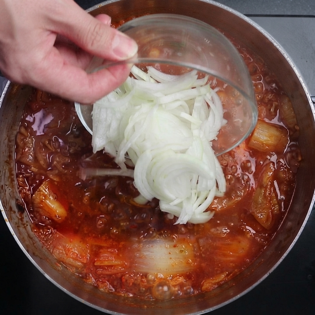 In Scheiben geschnittene weiße Zwiebeln werden aus einer Glasschüssel in eine kräftig kochende rote Kimchi-Brühe gegeben.
