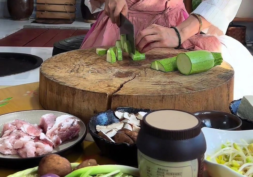 Hände schneiden eine grüne Zucchini mit einem Beil auf einem runden Holzschneidebrett in dreieckige Stücke.