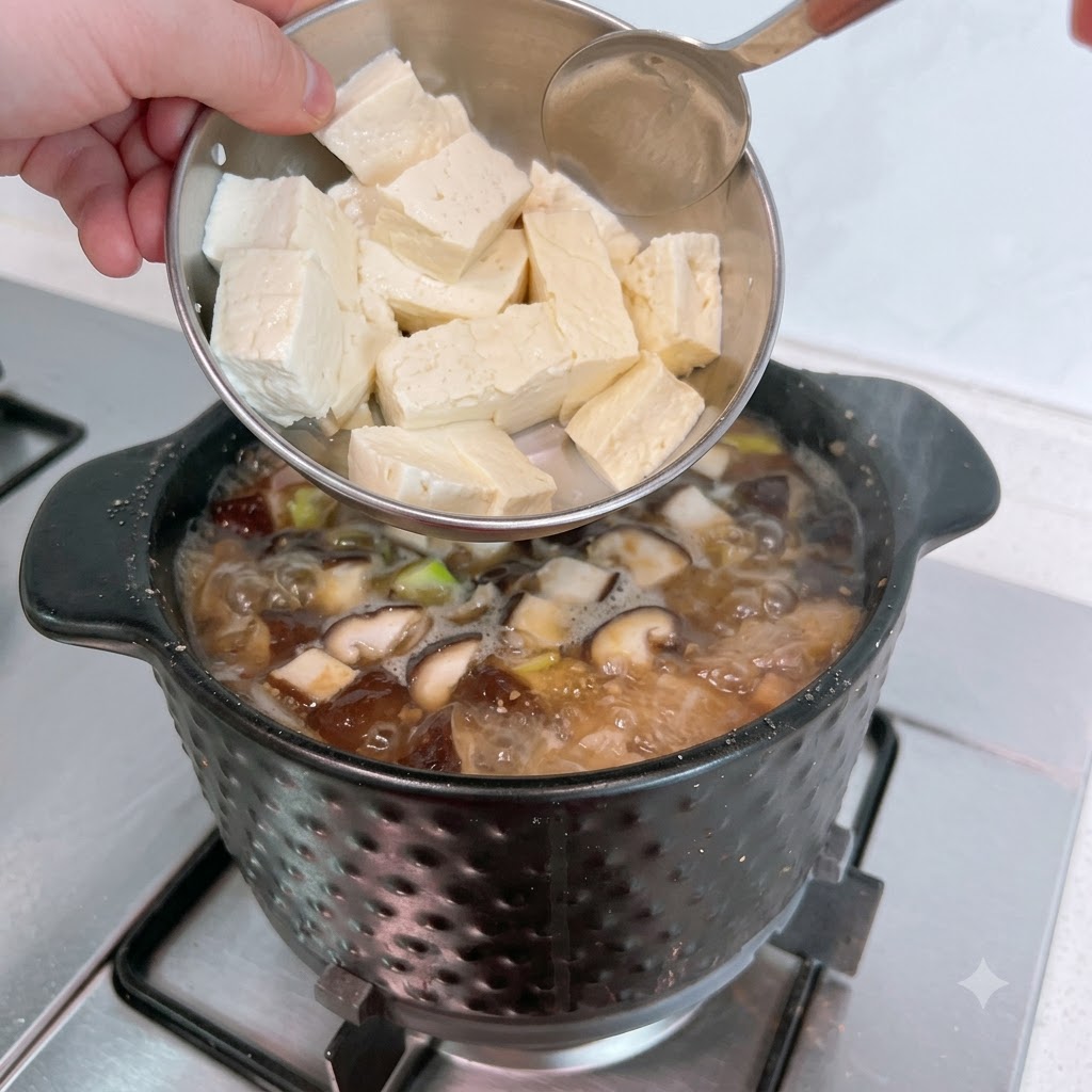 In Scheiben geschnittener weißer Tofu wird aus einer Metallschüssel in den blubbernden Eintopf gegeben.