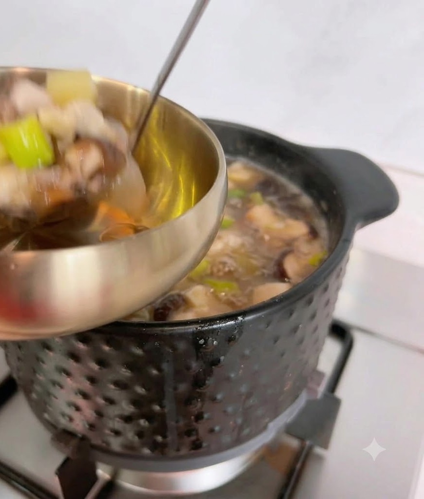Eine Schöpfkelle löffelt heiße Sojapastensuppe und Zutaten aus einem schwarzen Topf in eine metallene Servierschüssel.