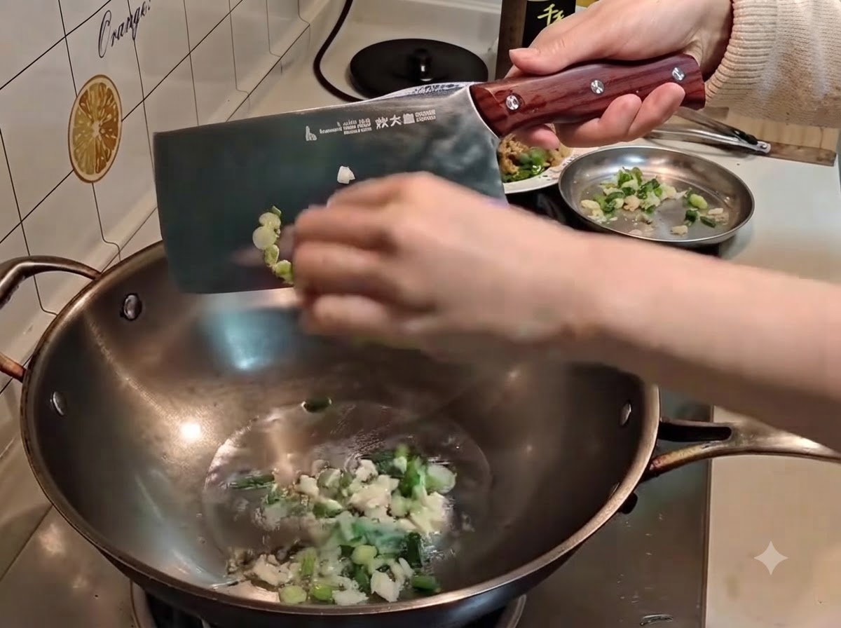 Eine Hand schiebt mit einem großen Metallbeil gehackte Frühlingszwiebeln und Knoblauch in das heiße Öl im Wok.