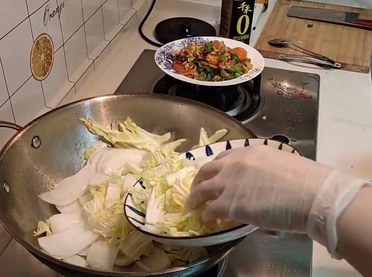 Eine handgeschützte Hand legt frisch geschnittenen rohen Chinakohl von einem gemusterten Teller in den Wok.