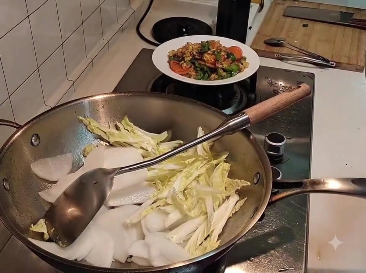 Geschnittener weißer Rettich liegt auf leicht eingefallenem Chinakohl in einem Metallwok, ein Pfannenwender liegt bereit.