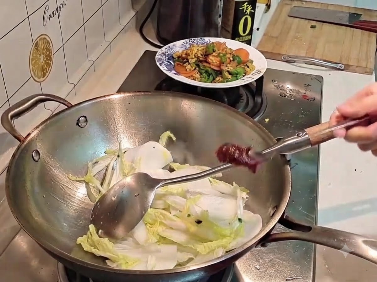 Eine Hand hält einen Löffel mit dunkler roter koreanischer Chilipaste über einen Wok voller Kohl und Rettich.