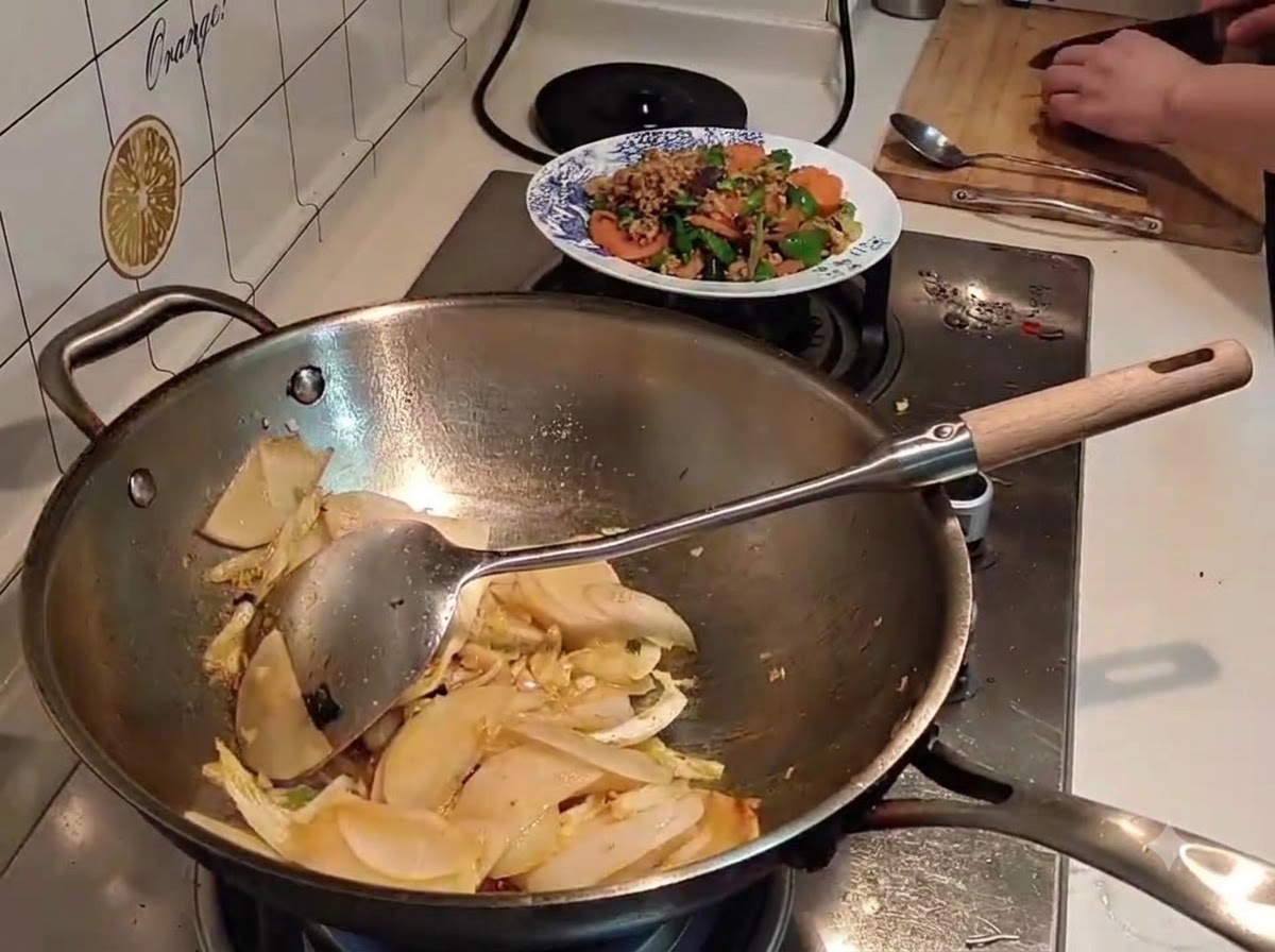 Wasser wird aus einem schwarzen Kessel in einen mit geschnittenem Rettich und Kohl gefüllten Wok gegossen.