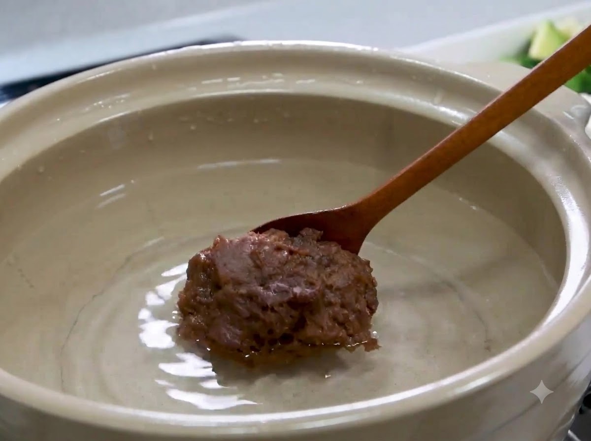 Ein Holzlöffel mit einem großen Klumpen koreanischer Sojabohnenpaste über dem Wasser in einem Topf.