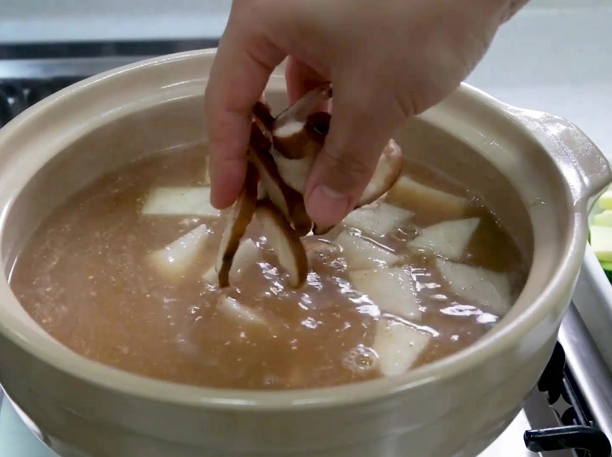Eine Hand gibt in Scheiben geschnittene Shiitake-Pilze in einen Topf mit Brühe und Rettichscheiben.