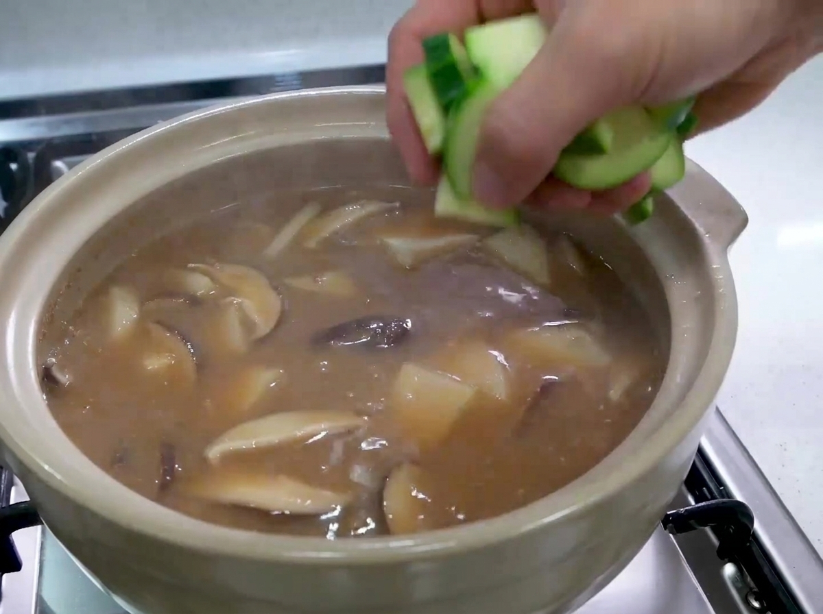 Eine Hand gibt gehackte Stücke roher grüner Zucchini in einen sprudelnden koreanischen Eintopf.