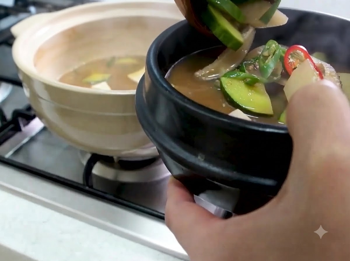 Eine Schöpfkelle füllt heißes Doenjang Jjigae mit Zucchini und Gemüse in eine schwarze Servierschüssel aus Stein.