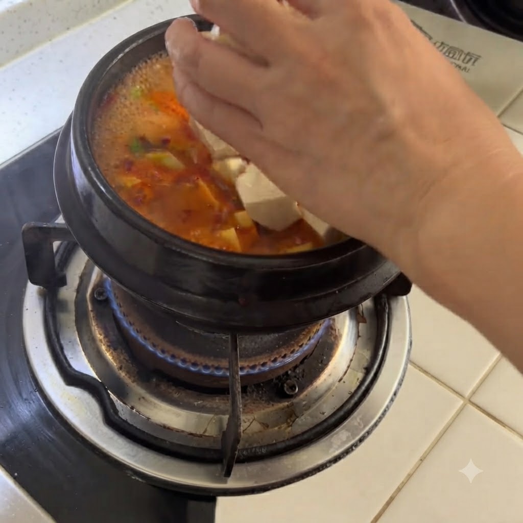Gewürfelter weißer Tofu wird von Hand in einen köchelnden Steintopf mit scharfer Suppe gegeben.