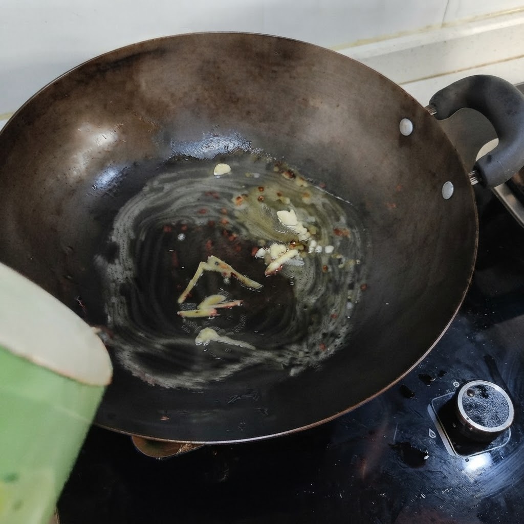 Kochendes Wasser wird aus einem grünen Behälter in einen Wok mit geschmolzenem Schmalz, Ingwer und Knoblauch gegossen.