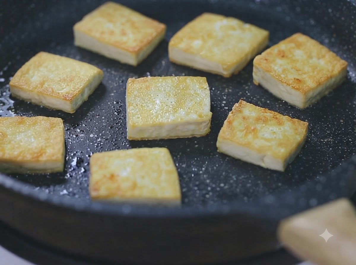 Mehrere goldbraune Tofuwürfel braten in einer schwarzen Pfanne bei niedriger Hitze an.