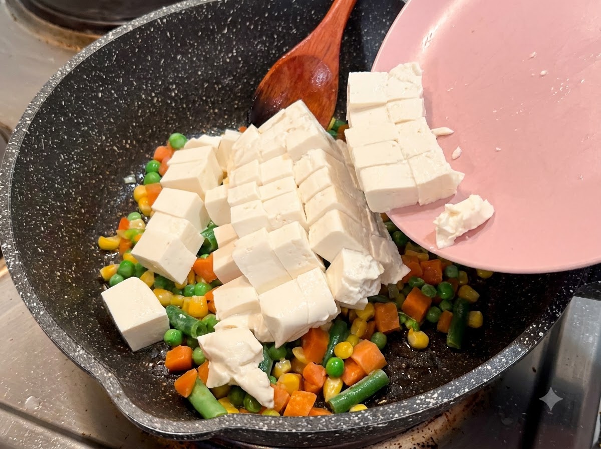 Gewürfelter weicher Tofu gleitet von einem rosa Teller in eine gesprenkelte Bratpfanne mit Gemüsemischung.
