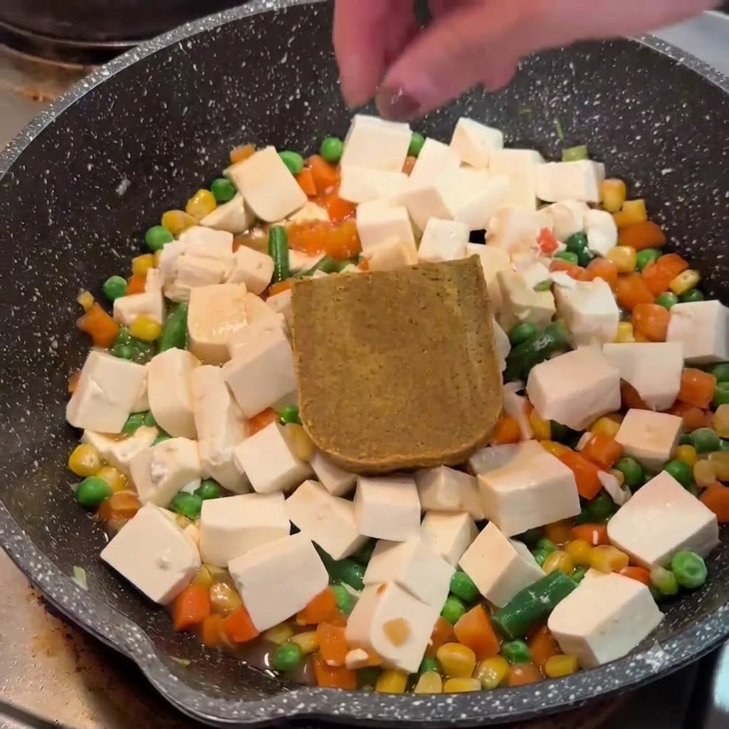 Eine Hand legt einen festen braunen Curry-Roux-Block auf Tofu und Gemüse in eine Pfanne.