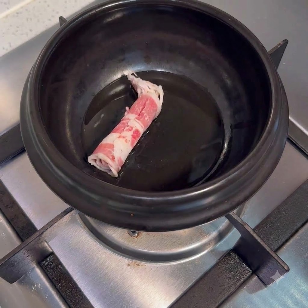 Rohe Rindfleischscheiben in einem dunklen Keramiktopf mit Öl auf einer Herdplatte.