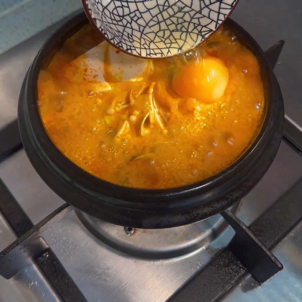 Ein rohes Ei wird aus einer kleinen Schüssel in den kochenden koreanischen Tofu-Eintopf geglitten.