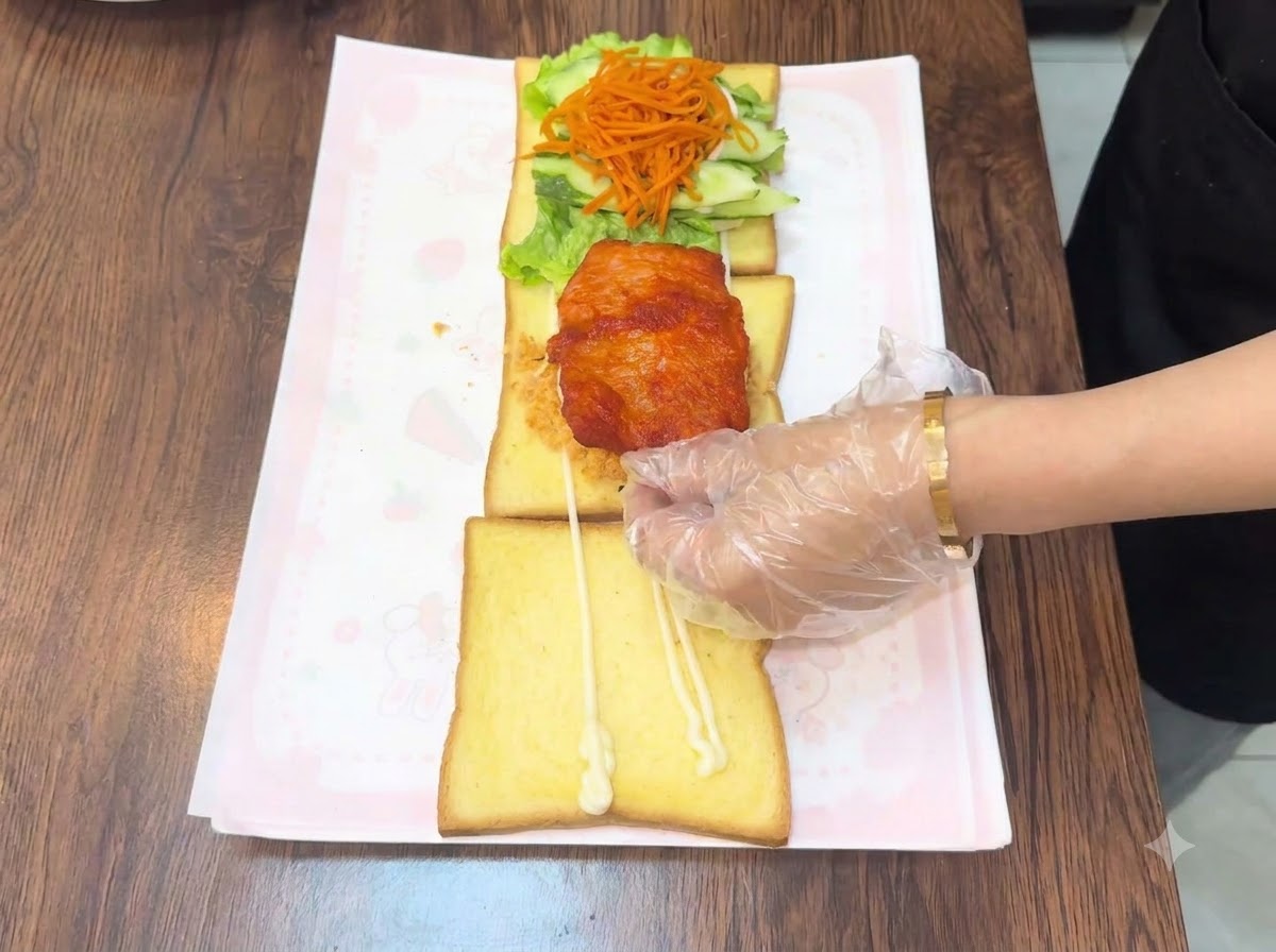 Eine handschuhbekleidete Hand platziert ein warmes, frittiertes Hähnchenschnitzel auf den geschichteten Toastaufbau.