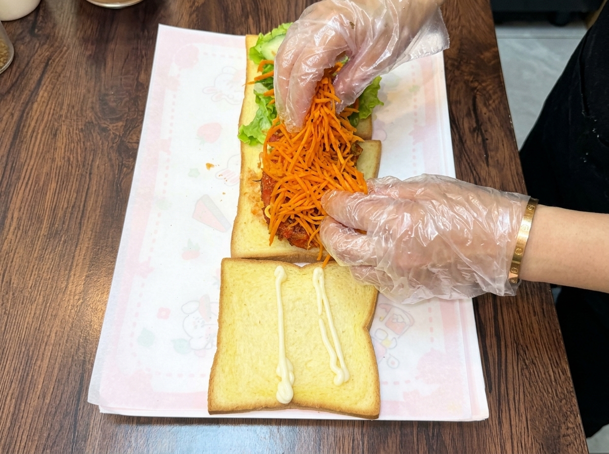 Handschuhbekleidete Hände häufen einen großzügigen Berg leuchtend orangefarbener, gebratener Karottenstreifen auf das gestapelte Hähnchenschnitzel.