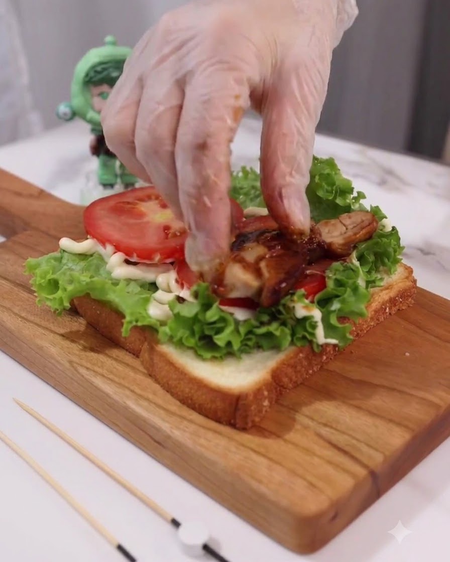 Eine mit Handschuhen bekleidete Hand legt gekochte Teriyaki-Hähnchenscheiben auf die Tomaten- und Salatschichten.
