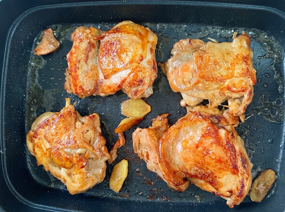 Vier Hähnchenkeulen braten in einer Pfanne mit goldbraun-knuspriger Haut und Ingwerscheiben.