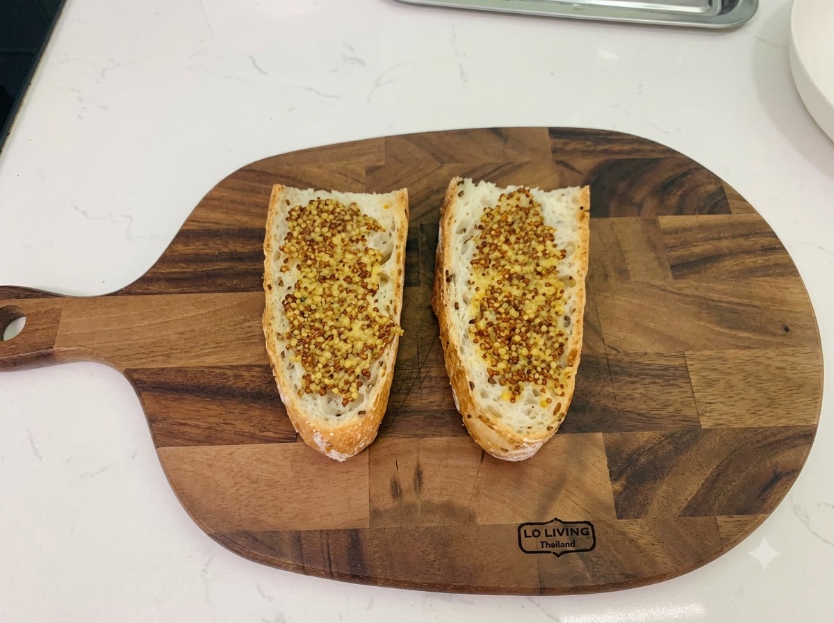 Zwei Scheiben Bauernbrot auf einem Holzschneidebrett, beide gleichmäßig mit körnigem Vollkornsenf bestrichen.