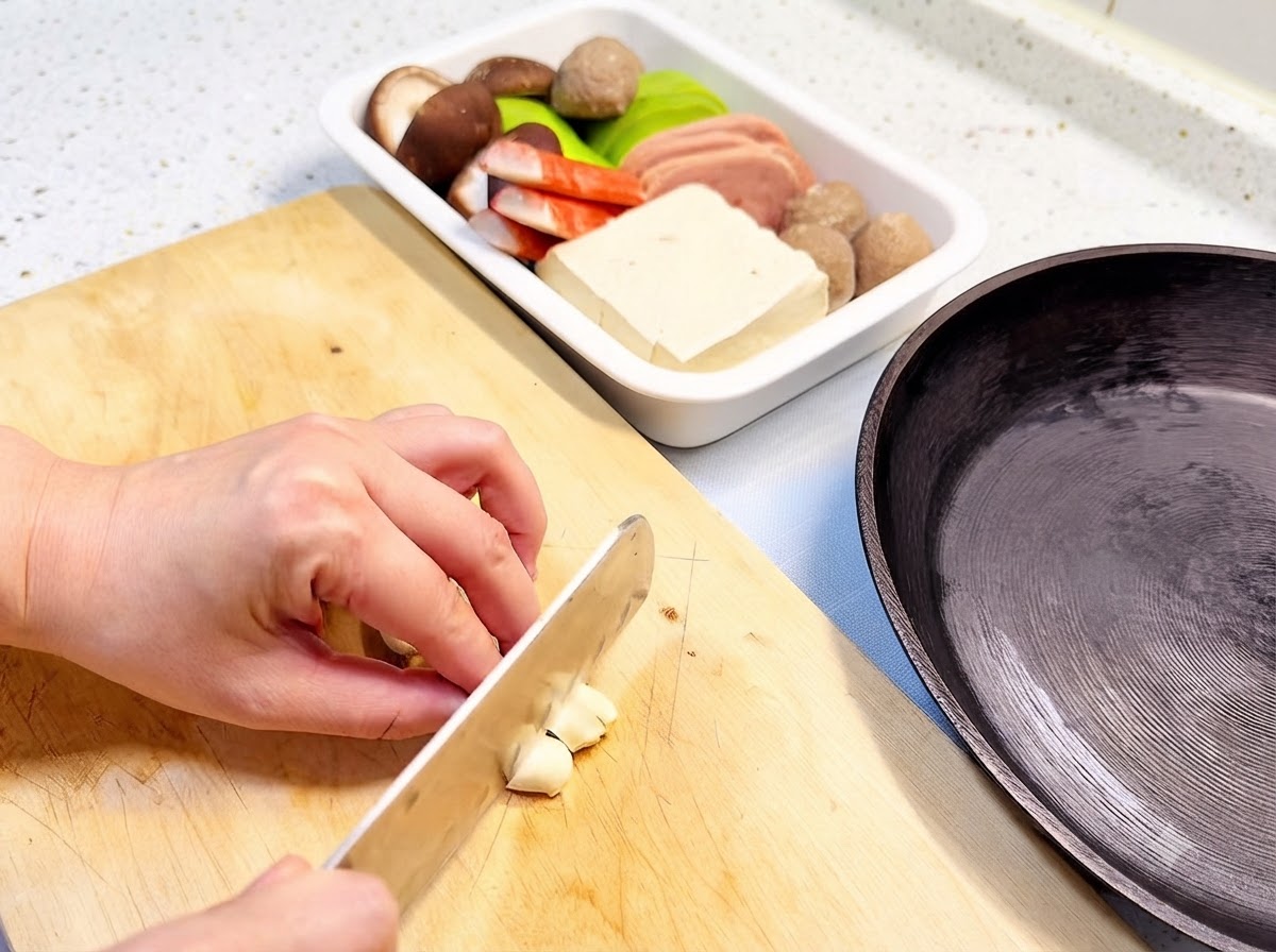 Knoblauchzehen werden auf einem Holzschneidebrett neben einer Bratpfanne und einer Zutatenschale in Scheiben geschnitten.