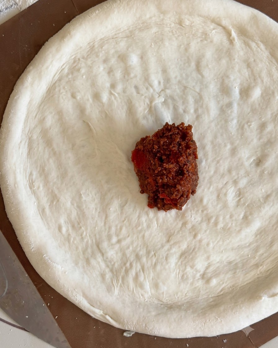 Ein kleiner Klecks dunkelroter Fleischsoße in der Mitte eines flachen Pizzateigkreises.