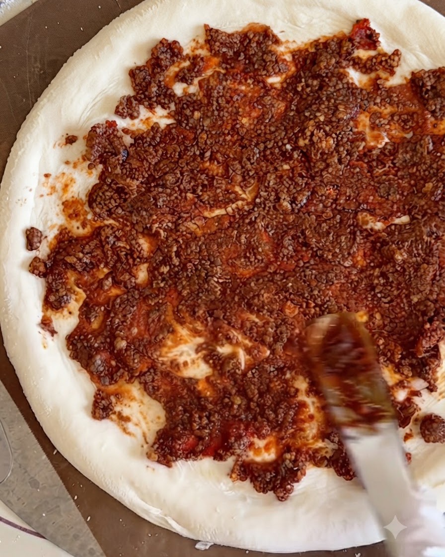 Gekochte Rindfleischwürfel werden über die rote Fleischsoße auf dem Pizzateig verteilt.