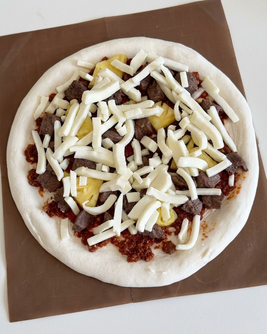 Geriebener weißer Mozzarella wird großzügig über die Rindfleisch- und Ananasbeläge gestreut.