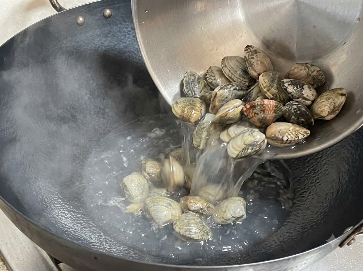 Frische Muscheln werden aus einer Metallschüssel in einen dampfenden Wok geschüttet.