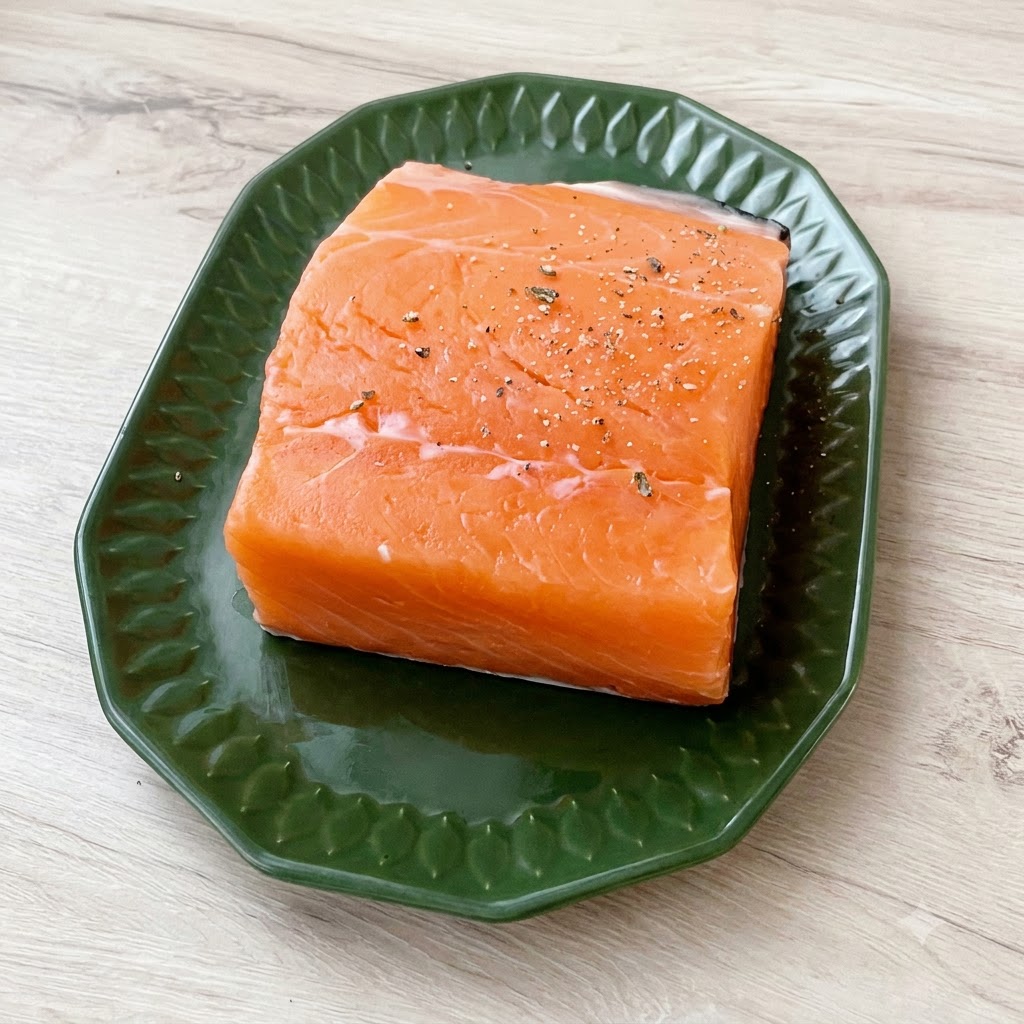 Ein dickes Stück frischer Lachs auf einem grünen Teller, großzügig bestreut mit grobem Meersalz und geschrotetem schwarzem Pfeffer.