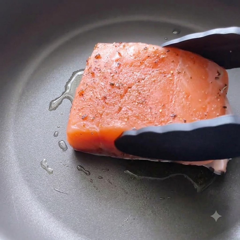 Ein gewürztes Lachsfilet wird mit einer schwarzen Küchenzange mit der Hautseite nach unten in eine geölte Pfanne gelegt.