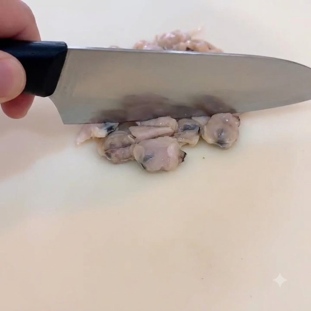 Nahaufnahme einer Hand, die mit einem Chefmesser gekochtes Muschelfleisch auf einem weißen Schneidebrett in kleine Stücke schneidet.