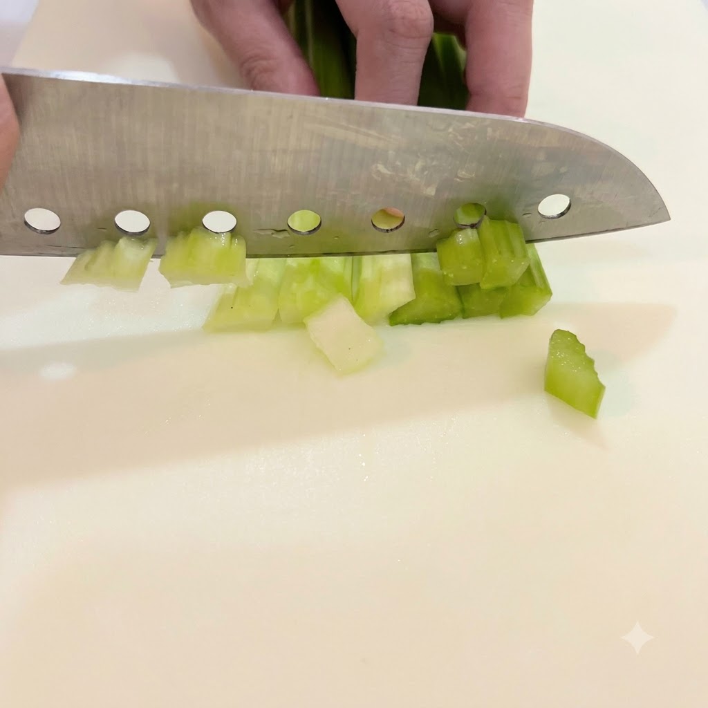 Eine Hand führt ein großes Messer, um hellgrüne Selleriestangen auf einem weißen Schneidebrett in kleine Stücke zu schneiden.