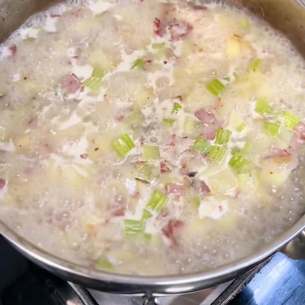 Milch und Schlagsahne werden in einen köchelnden Suppentopf mit sichtbaren Kartoffel-, Sellerie- und Speckstückchen gegossen.