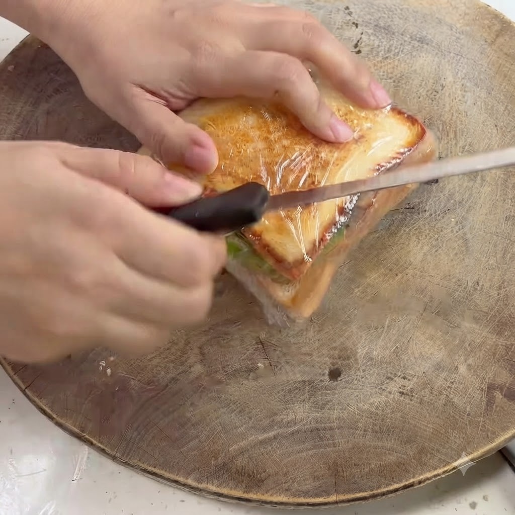 Ein Küchenmesser schneidet diagonal durch ein in Frischhaltefolie gewickeltes Sandwich auf einem Holzbrett.