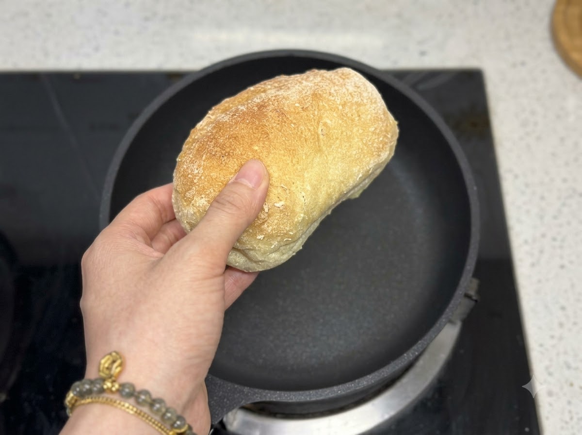 Eine Hand hält einen Laib Ciabattabrot direkt über eine leere schwarze Bratpfanne.