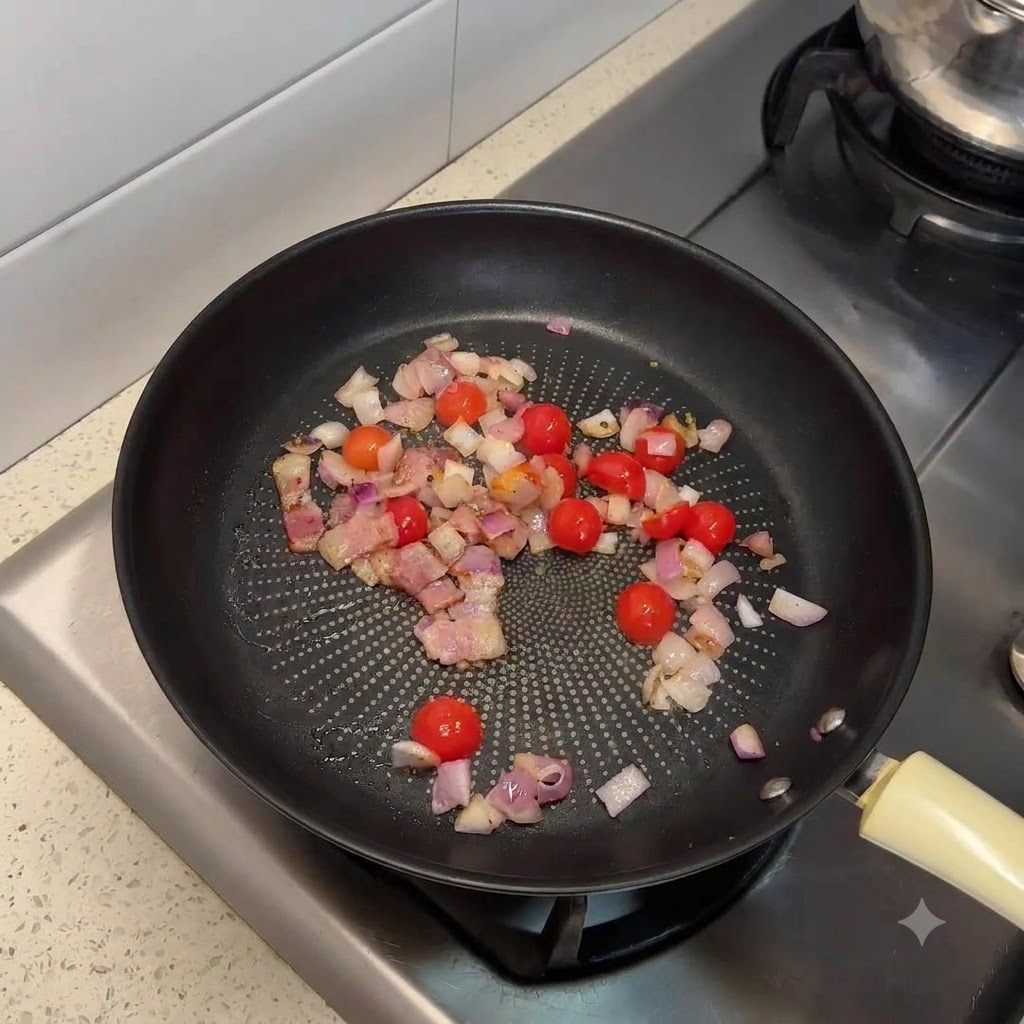 Eine Bratpfanne gefüllt mit angebratenem Bacon, gehackten Zwiebeln und halbierten Kirschtomaten.