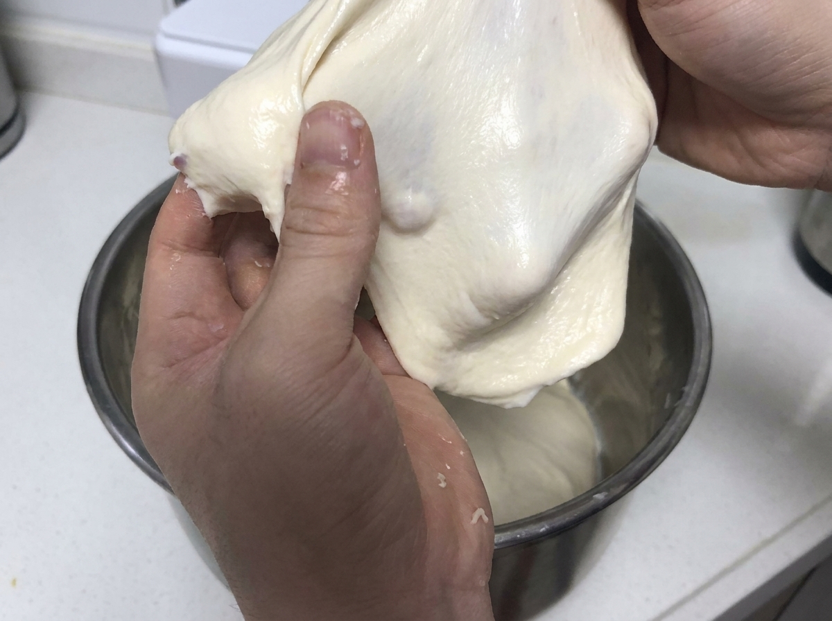 Hände ziehen und dehnen ein weiches, elastisches Stück weißen Teigs zur Demonstration der Glutenstärke.