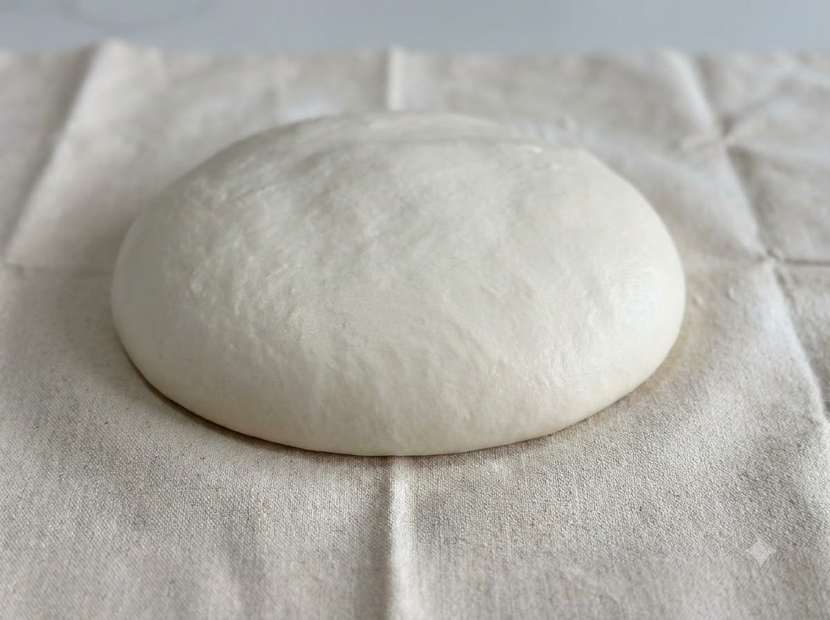 Eine glatte, gerundete Kuppel aus weißem Teig ruht auf einer bemehlten Stoffoberfläche.